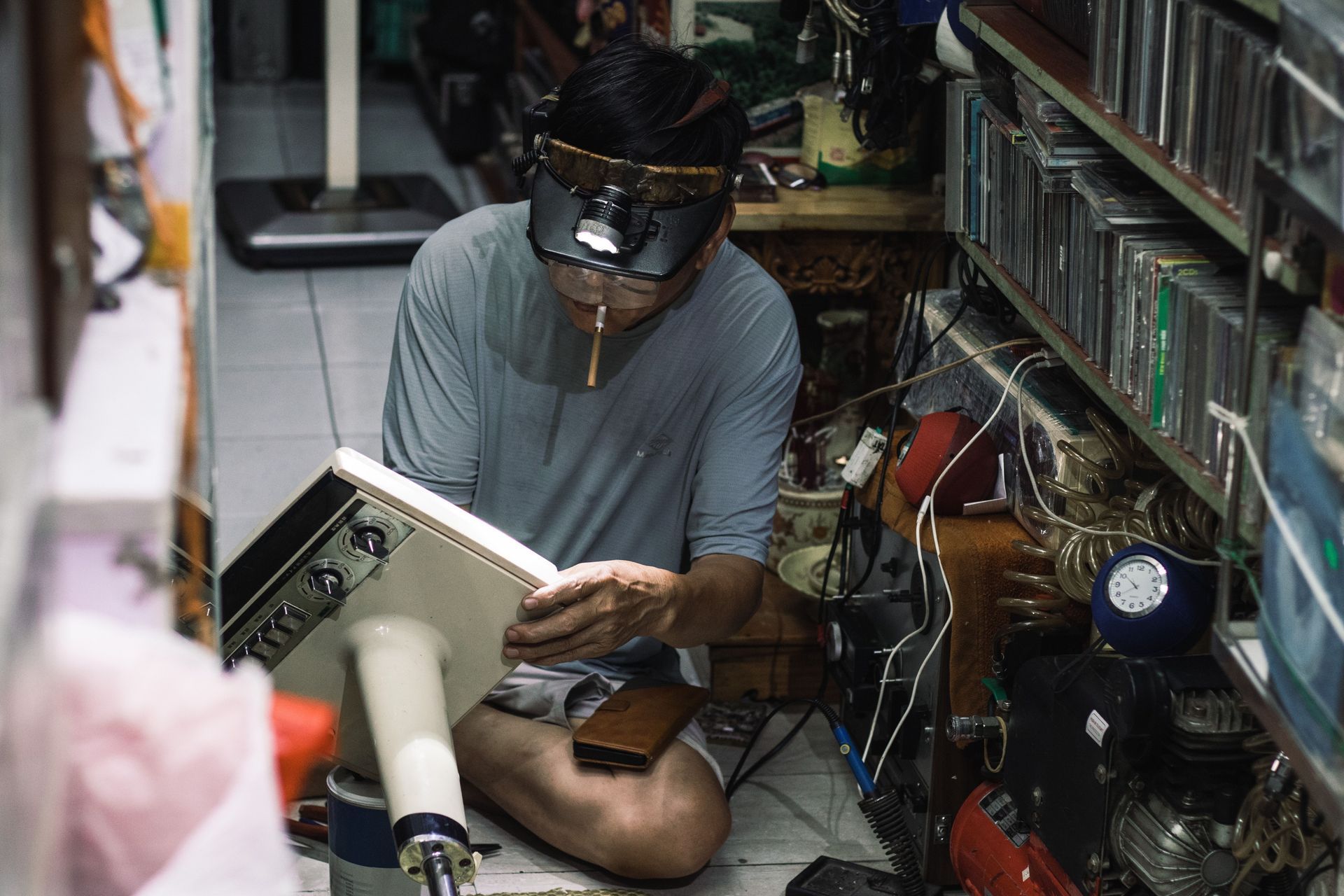 Person sitting on the floor in a cluttered room, reading a book under a black fan hat.
