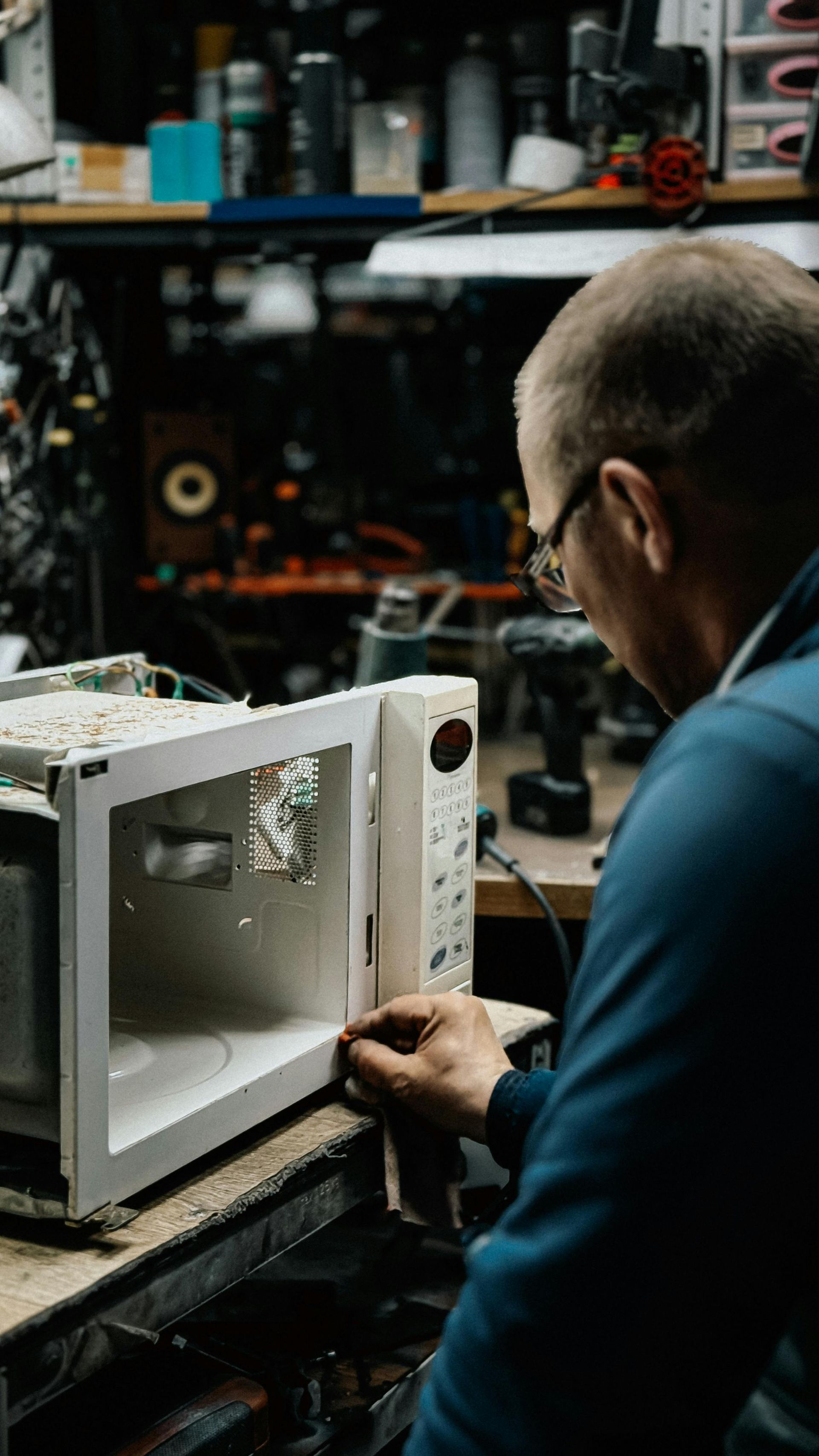 Technician repairing a microwave on a cluttered workshop bench, viewed from behind.