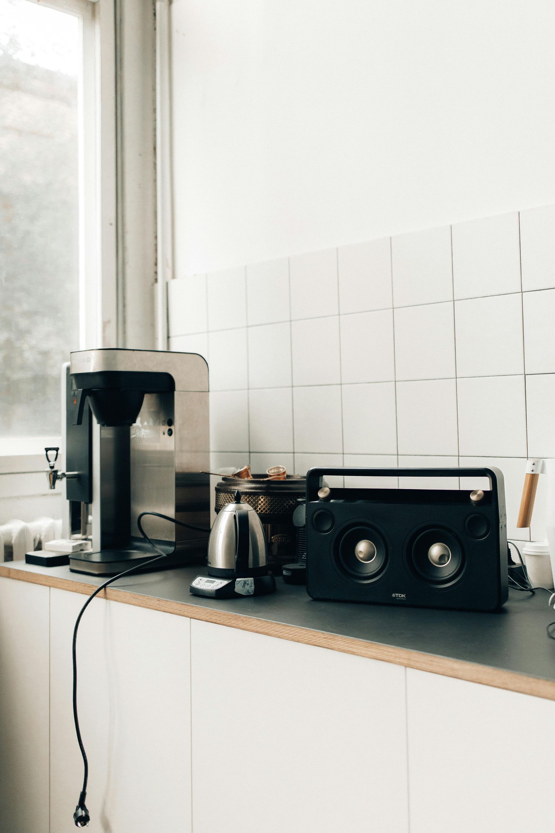Coffee maker, kettle, and speaker on a kitchen counter next to a window.