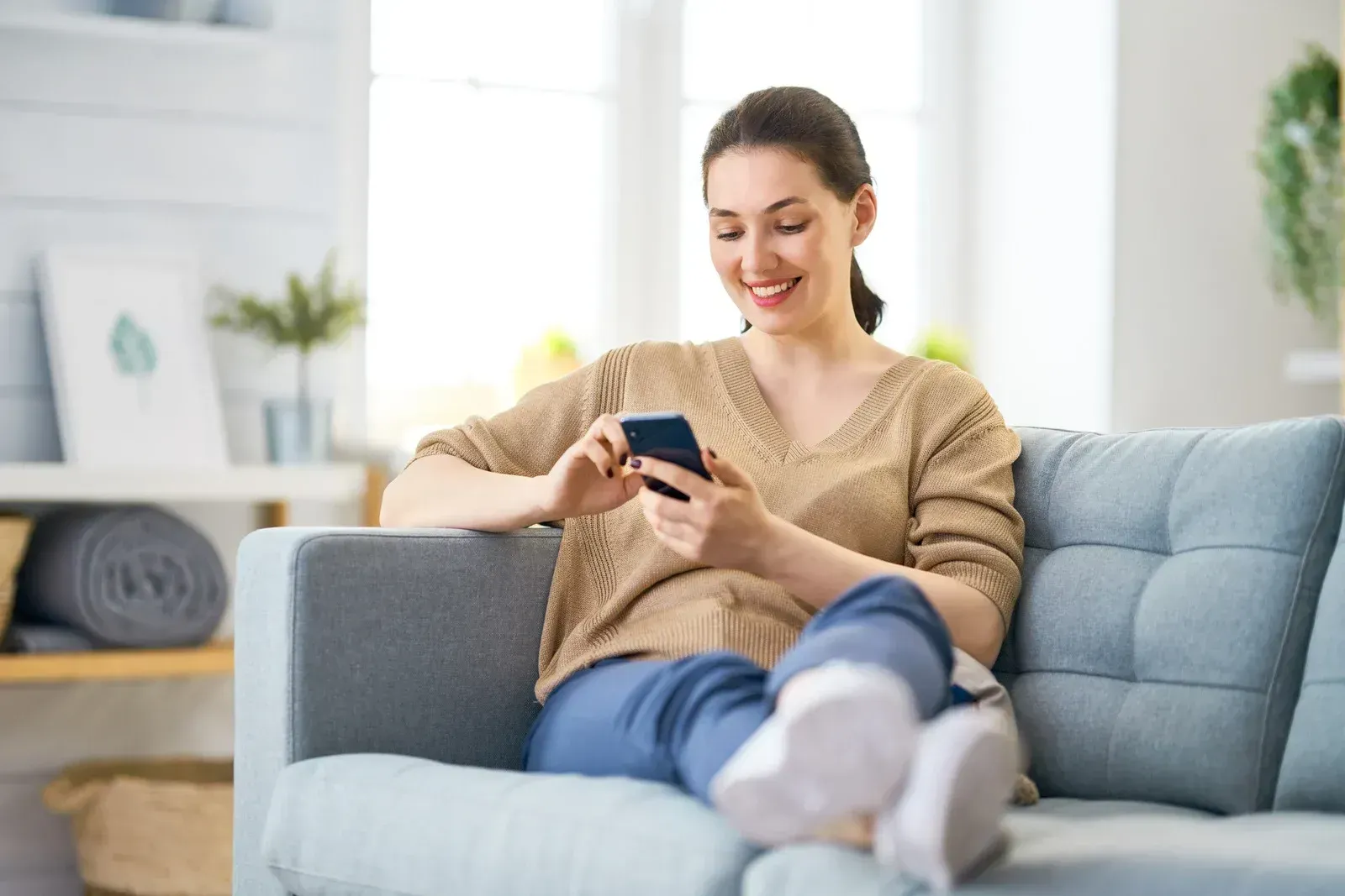 Woman relaxing on a light blue sofa, smiling at her phone in a bright living room.
