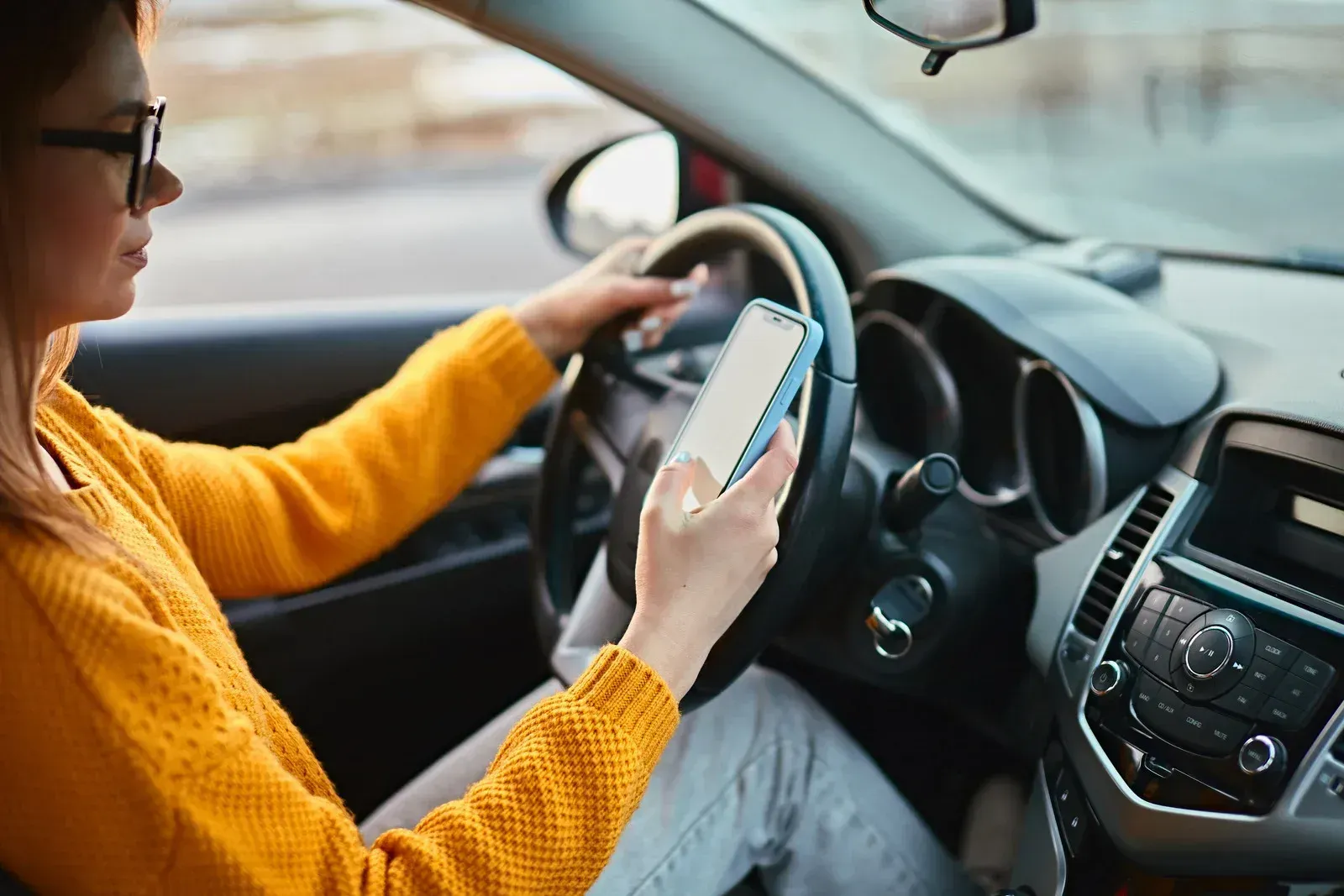 Person in orange sweater driving while looking at a phone inside a car