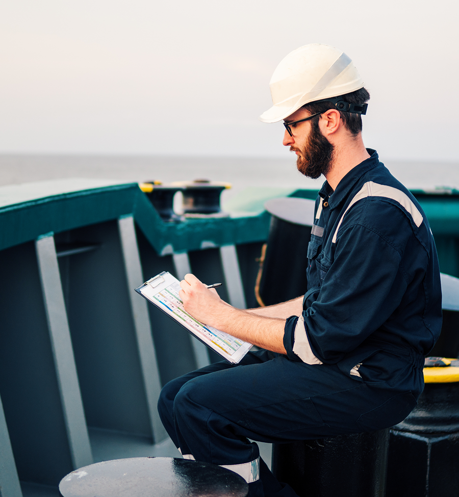 A man wearing a hard hat is sitting on a boat writing on a clipboard.