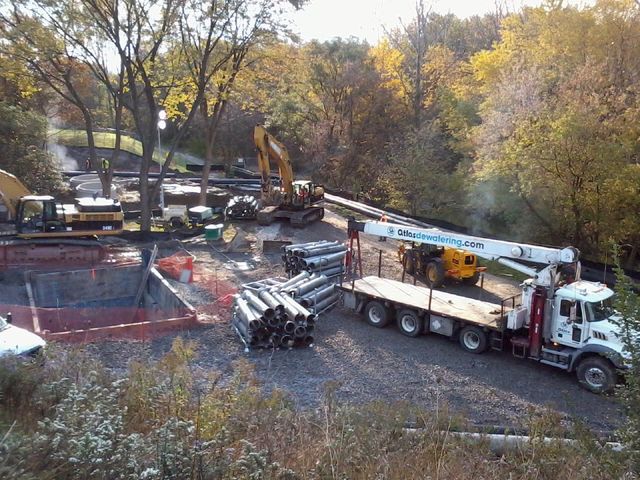 Construction site with heavy machinery and pipe bundles, autumn foliage in the background.