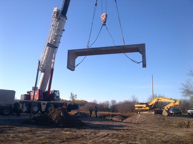 A crane lifts a concrete bridge segment above a construction site.