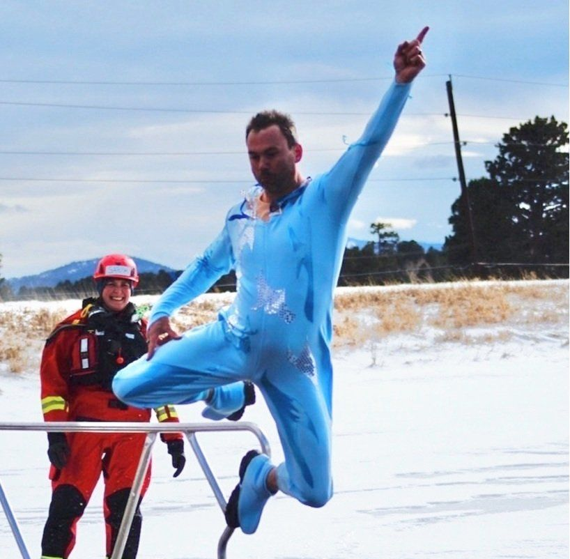Man in blue jumpsuit jumps over railing, pointing up. Snow and a person in red suit in the background.