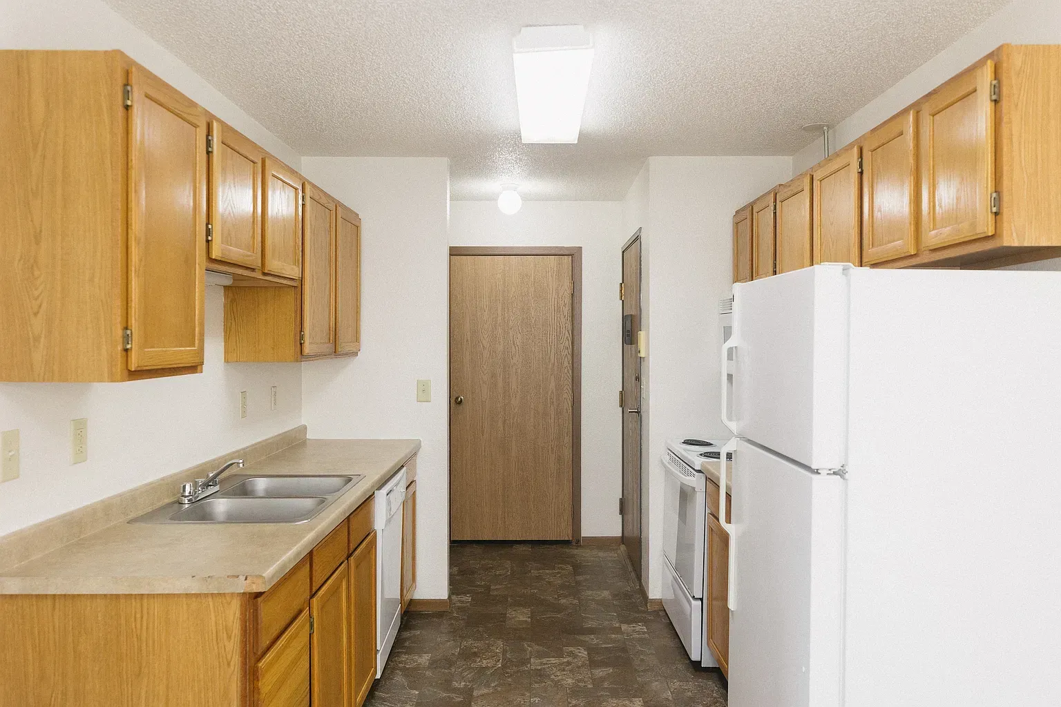 Kitchen with wooden cabinets, beige countertops, white appliances, and a hallway with a door.