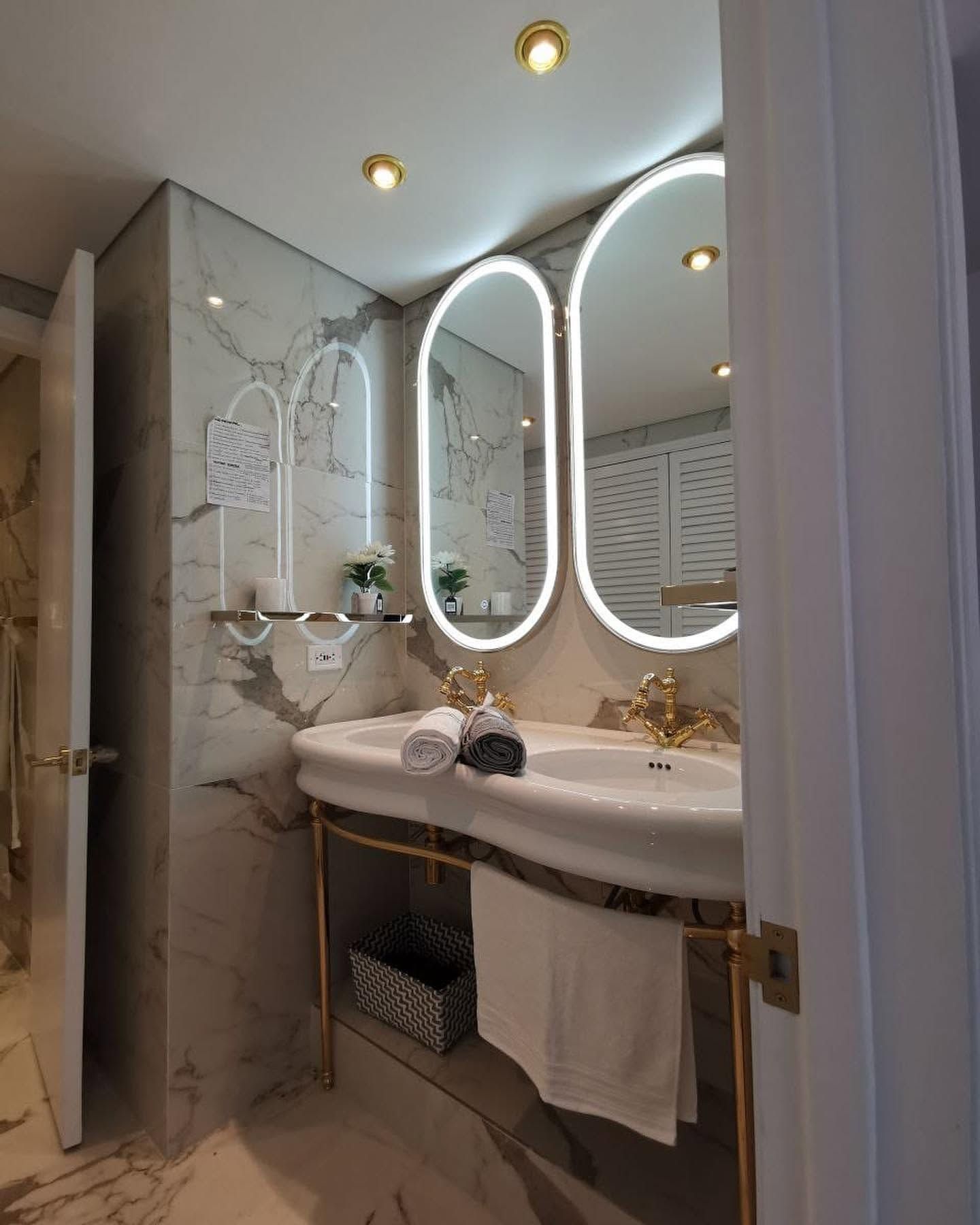 Elegante baño con dos espejos ovalados, lavabo blanco, grifería de latón e iluminación tenue.