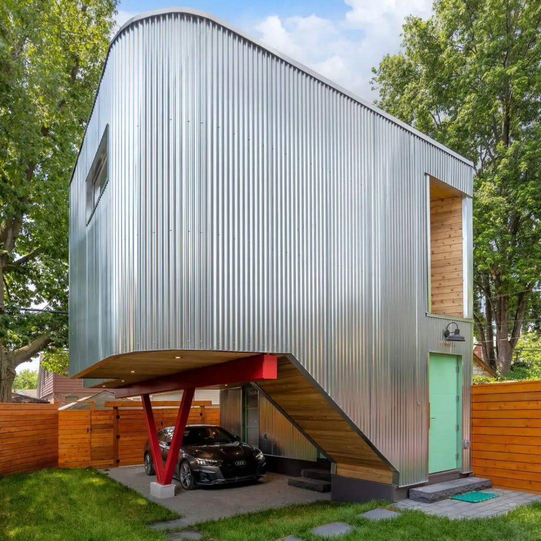 Moderna casa revestida de plata sobre pilotes con cochera, puerta verde y valla de madera en un patio frondoso.