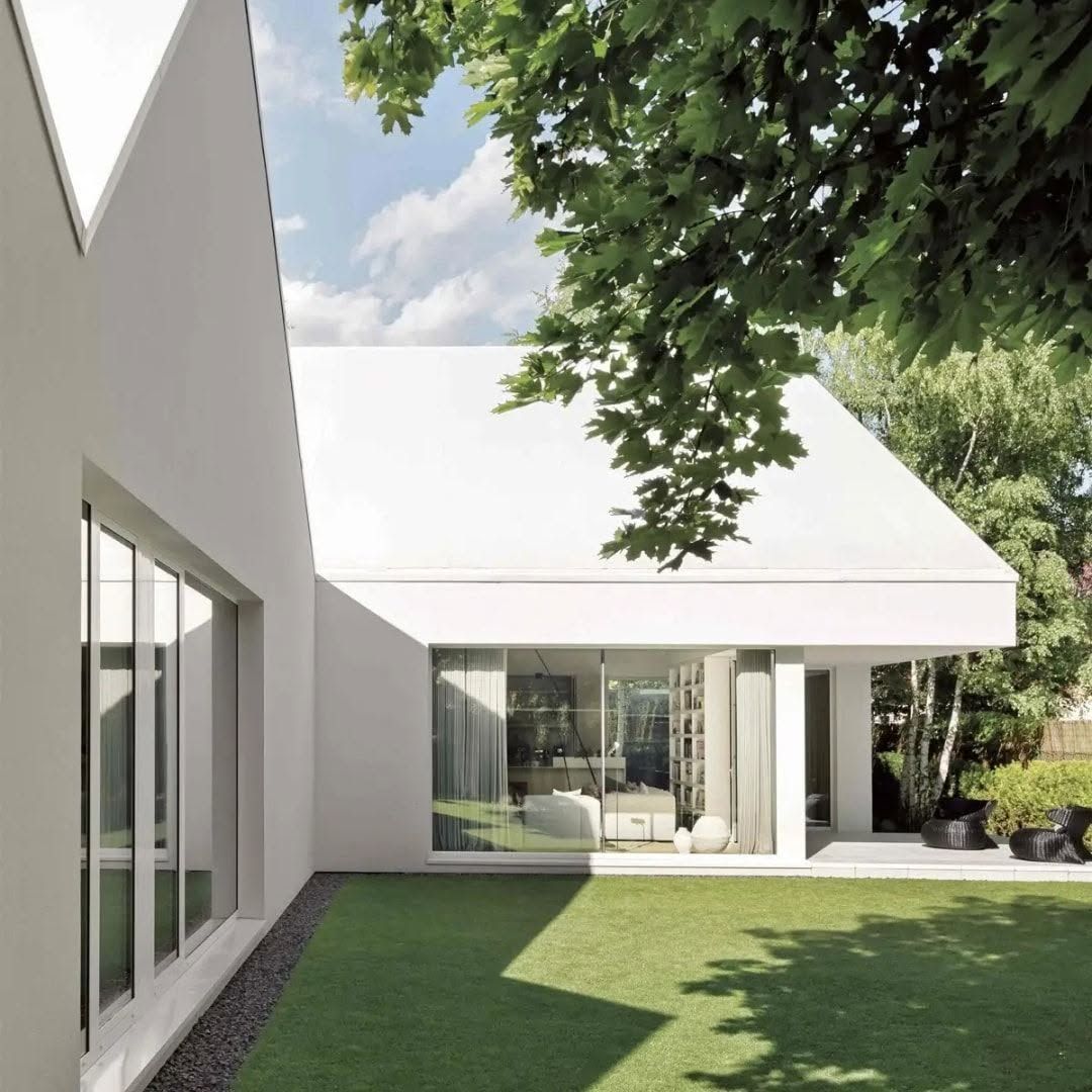 Casa blanca moderna con grandes ventanales y un césped verde, a la sombra de los árboles.