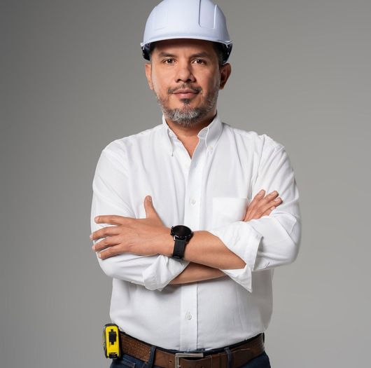 Obrero con casco y camisa blancos, con los brazos cruzados, de pie sobre un fondo gris.