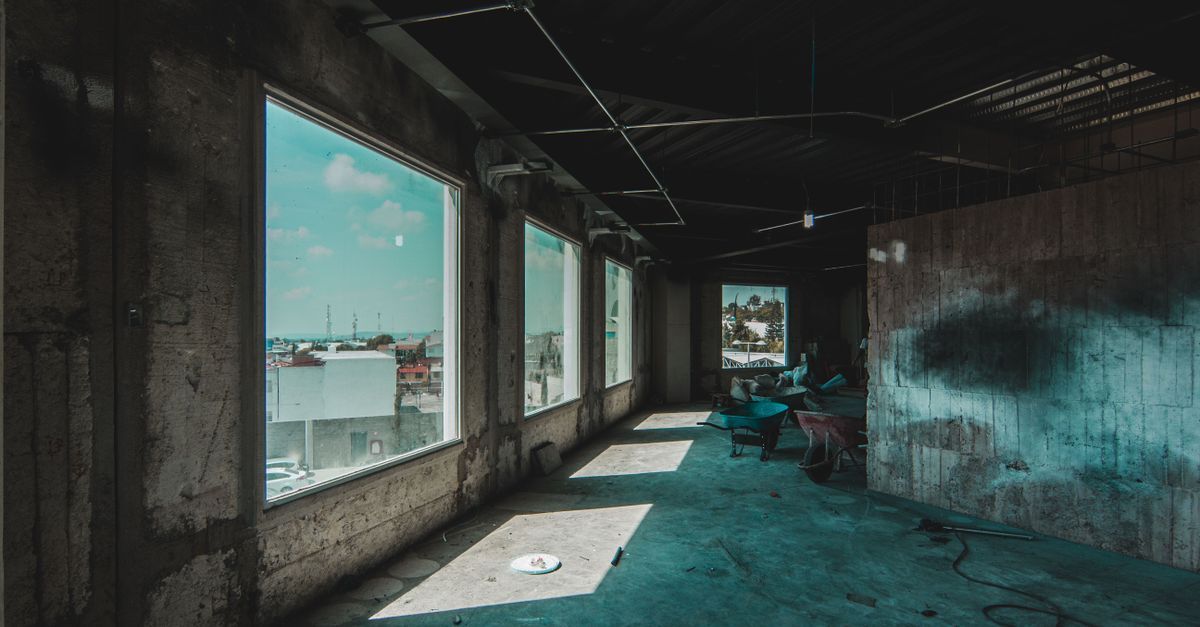 Pasillo industrial abandonado con grandes ventanales, suelo turquesa y luz solar que proyecta largas sombras.