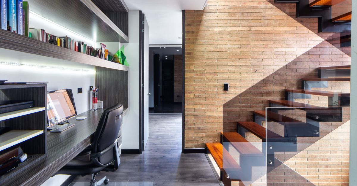 Oficina moderna en casa con escritorio, silla, estantes de pared y escalera de madera junto a una pared de ladrillo.