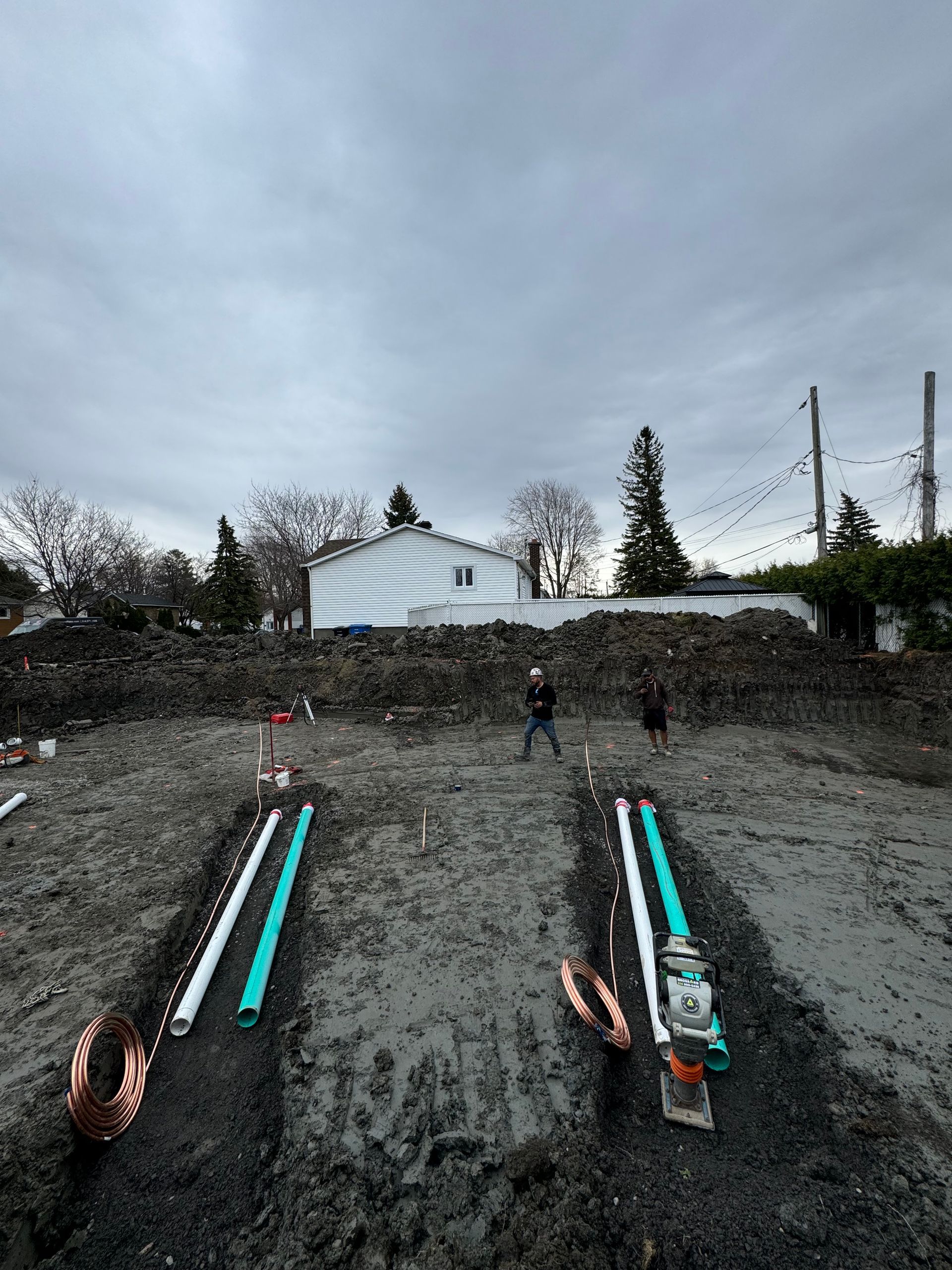 Un chantier avec beaucoup de tuyaux dans la terre et une maison en arrière-plan.