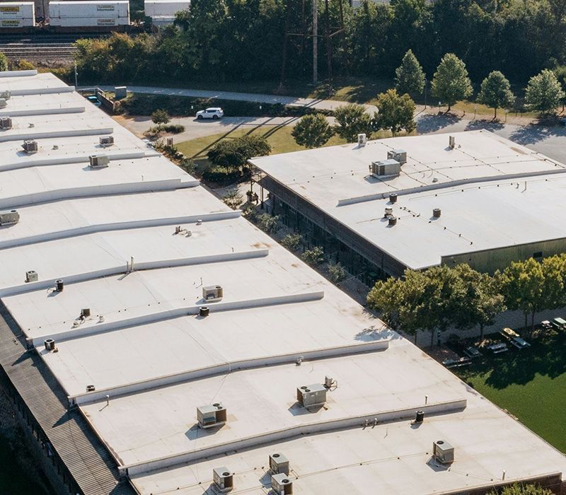 Aerial view of two flat-roofed industrial buildings featuring multiple HVAC units, surrounded by trees and paved parking.