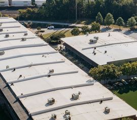 Aerial view of two commercial warehouse buildings with light-colored flat roofs, HVAC units, and surrounding trees.