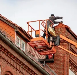 A construction worker in a raised orange lift works on the roof of a brick building.
