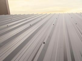 A low-angle view of a grey metal roof with repeating corrugated patterns and visible screw fasteners under a pale sky.