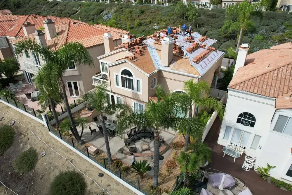 An aerial view of a residential house under construction, with workers on the roof replacing clay tiles.