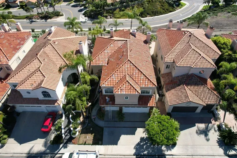 Aerial view of three suburban homes with red tile roofs, each featuring a garage and driveway, on a sunlit street.