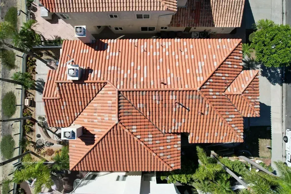 An aerial view of a terra-cotta tile roof on a residential home with two white chimneys and surrounding greenery.