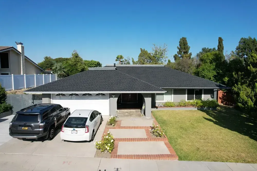 A single-story gray house with a black shingled roof, a two-car garage, and a front yard with a concrete walkway.