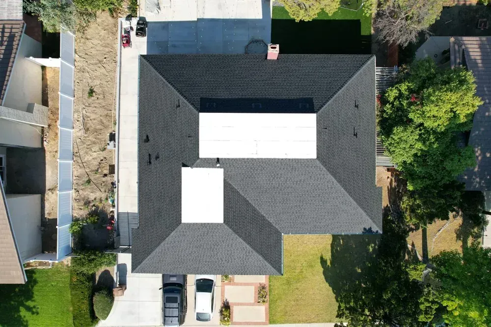 A top-down aerial view of a dark shingled house roof, featuring two white rectangular areas, set among trees and a driveway.
