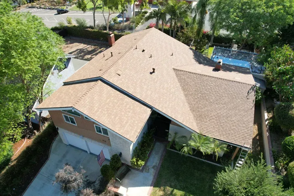 An aerial view of a single-story suburban house with a brown shingled roof, a garage, and a swimming pool in the backyard.