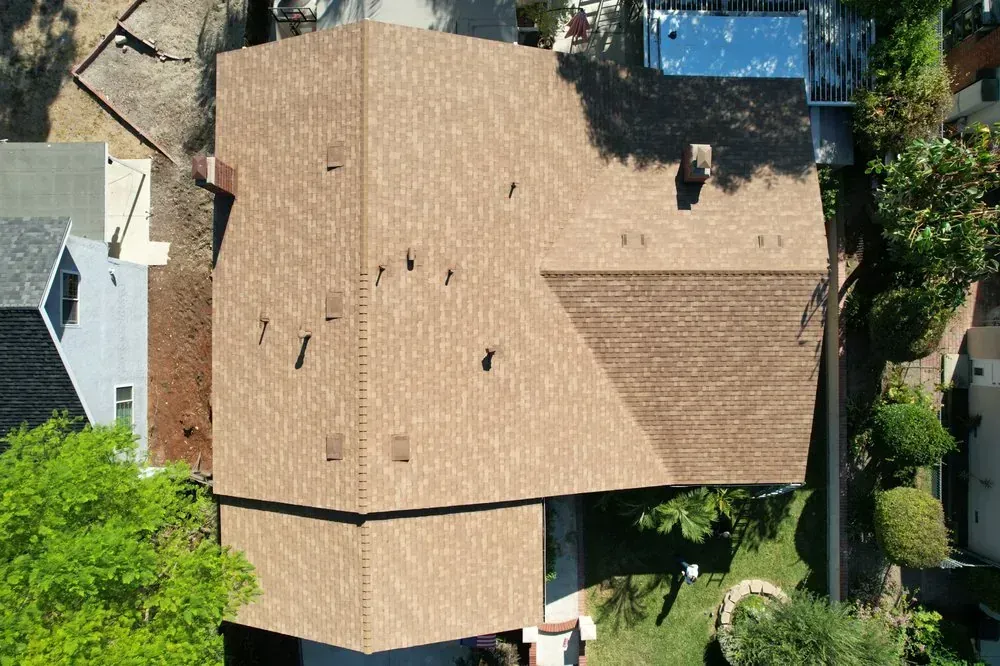 An aerial view of a brown asphalt-shingled house roof, surrounded by trees and neighboring yards.