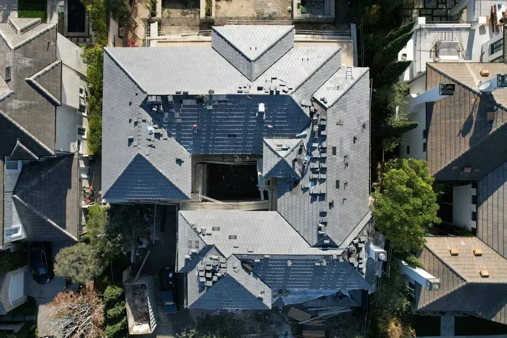 Aerial view of a residential house with a complex gray shingle roof, featuring a large central open courtyard area.