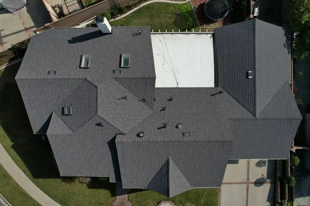 An aerial view of a dark grey shingled residential roof with a rectangular flat section and multiple skylights.