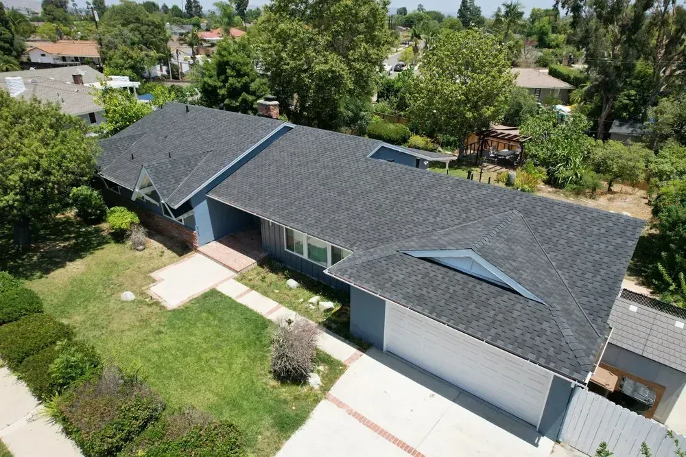 An aerial view of a blue single-story house with a grey shingled roof, a two-car garage, and a front lawn.