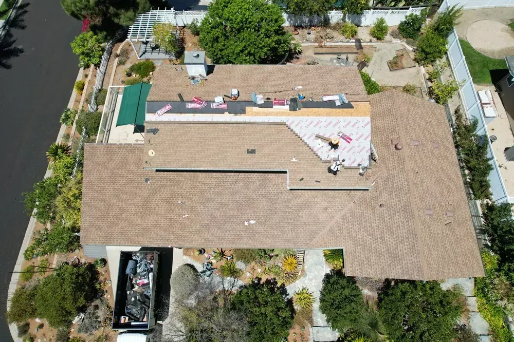 Aerial view of a residential roof undergoing repairs with workers, construction materials, and a dumpster on site.