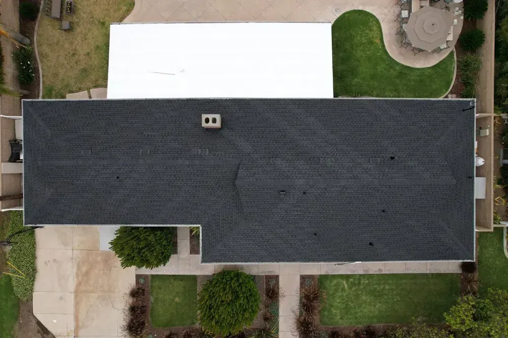 An aerial view of a dark shingled residential roof with a white rectangular patio cover below it, surrounded by lawns.