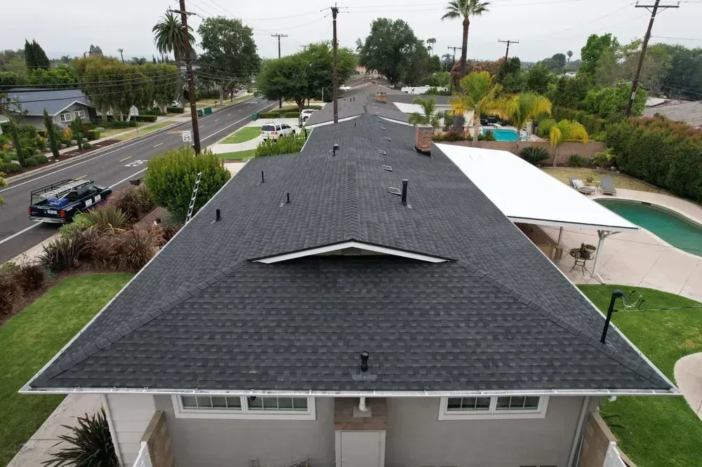 An aerial view of a dark-shingled residential roof, with a swimming pool and suburban street in the background.