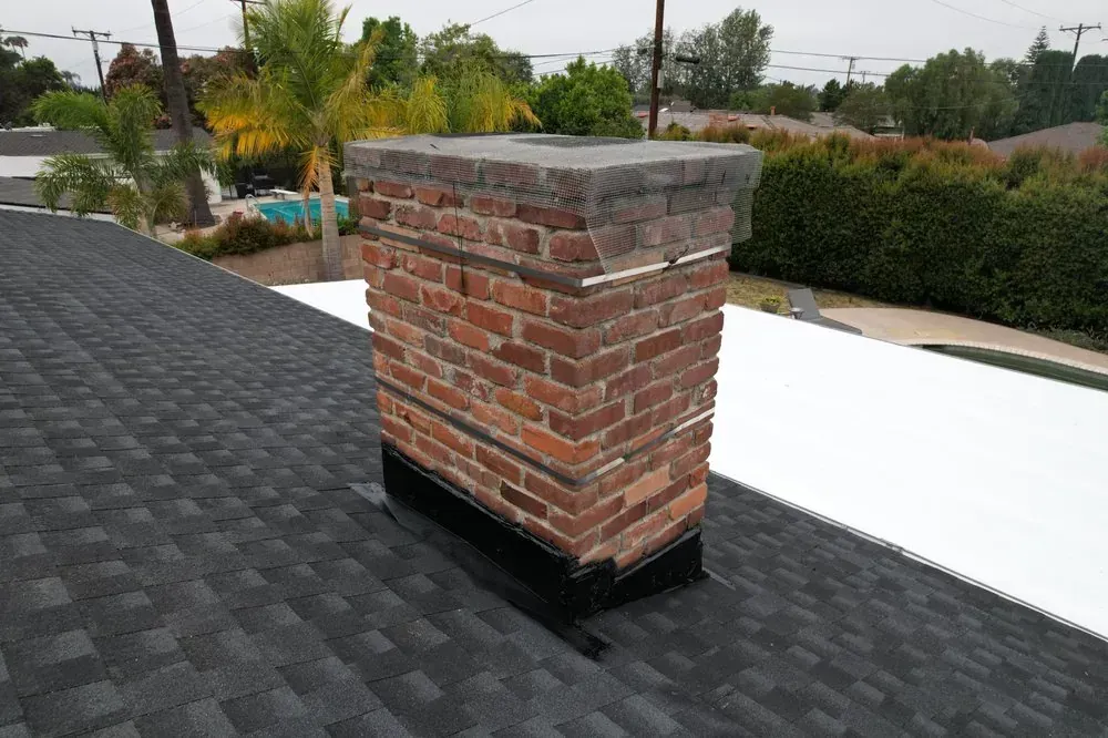 A brick chimney rises from a roof featuring both dark asphalt shingles and a section of white flat roofing.