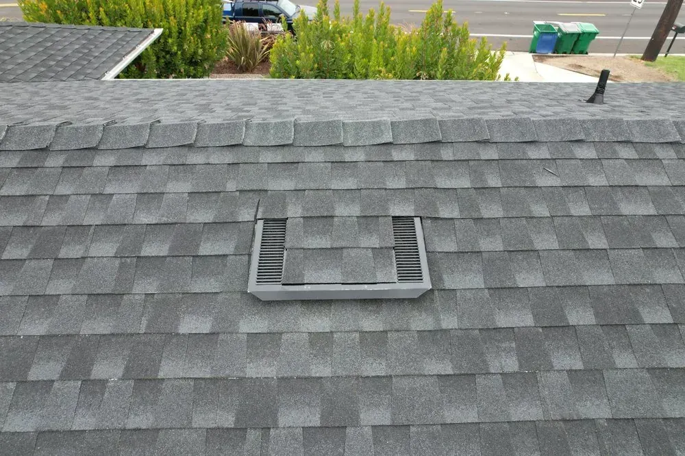 A low-profile roof vent installed on a gray shingled roof, viewed from an elevated position.