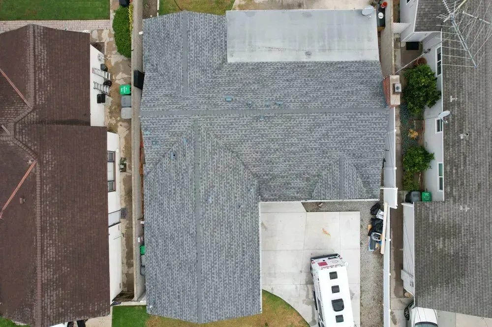 A bird's-eye view of a gray shingled roof on a residential house situated between neighboring homes and a driveway.