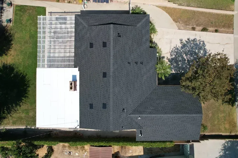 An aerial top-down view of a house roof featuring dark grey shingles and a white flat roof section, surrounded by trees.