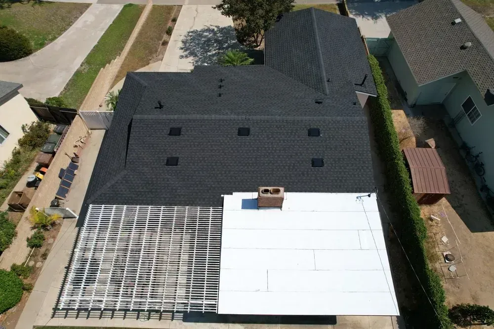 Aerial view of a house roof featuring a large black shingled section, a white flat section, and a lattice patio cover.