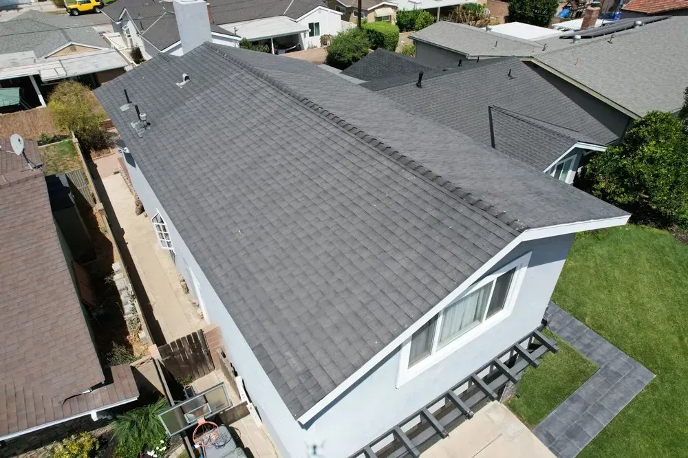 A high-angle view of a gray shingled roof on a house with a white exterior, located in a suburban neighborhood.