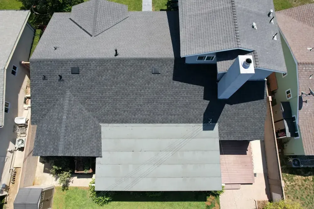 Aerial view of a residential shingled roof with a flat, light-colored patio cover in the backyard.