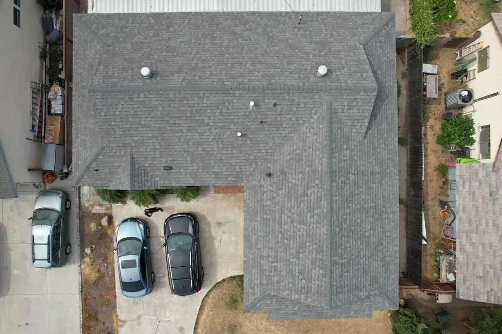 An aerial view of a gray shingled roof on an L-shaped house, with three parked cars on the concrete driveway below.