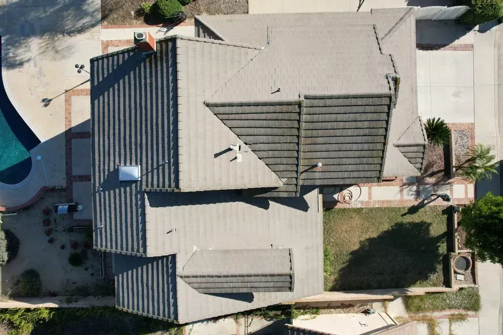 A top-down aerial view of a gray tiled residential roof with a chimney and skylight, surrounded by a yard and pool.