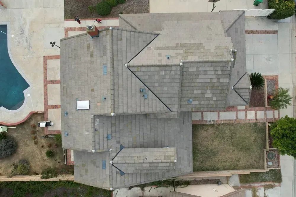 An aerial view of a gray-tiled house roof featuring multiple gables, a chimney, and a small skylight.