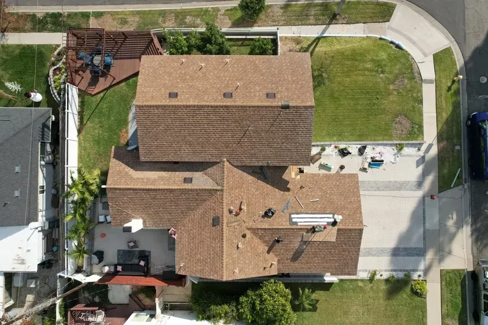 Aerial view of a residential home with a tan shingled roof, a paved driveway, and backyard landscaping.