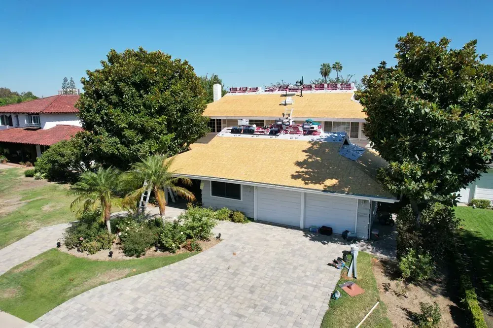 A two-story suburban house with a partially re-shingled yellow roof, a paved driveway, and large trees on a sunny day.
