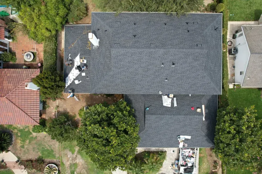 An aerial view of a dark grey shingled roof under construction, featuring workers, equipment, and a dumpster below.