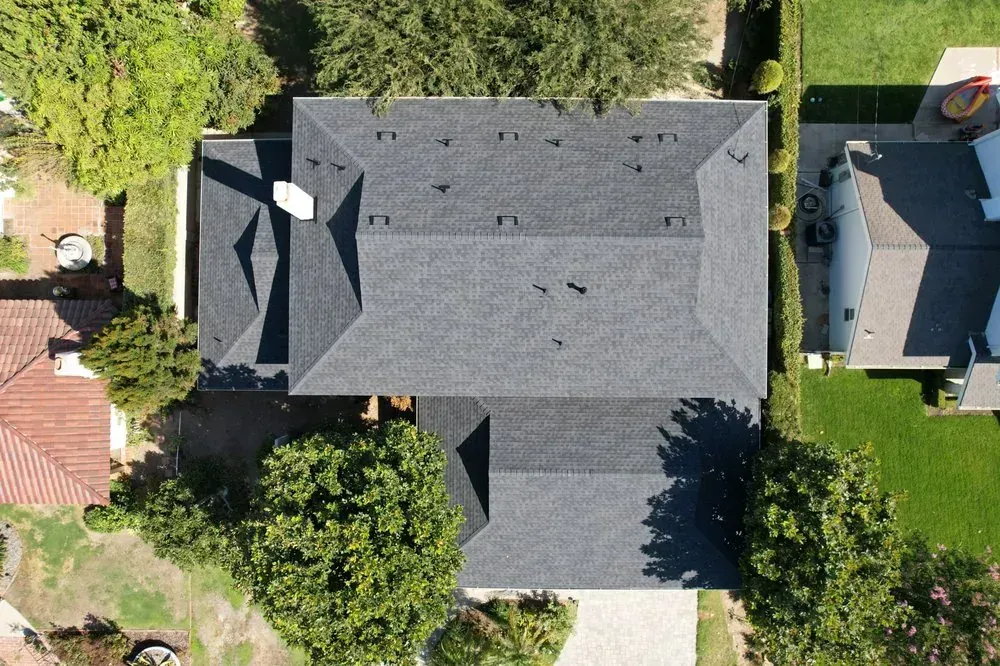 An aerial, top-down view of a dark-shingled residential roof surrounded by green trees and adjacent neighboring houses.