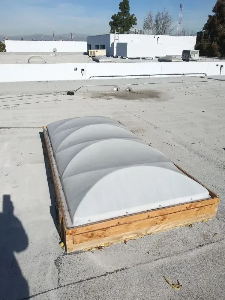 A rectangular, arched plastic skylight set in a wooden frame on a flat, light-colored commercial roof.