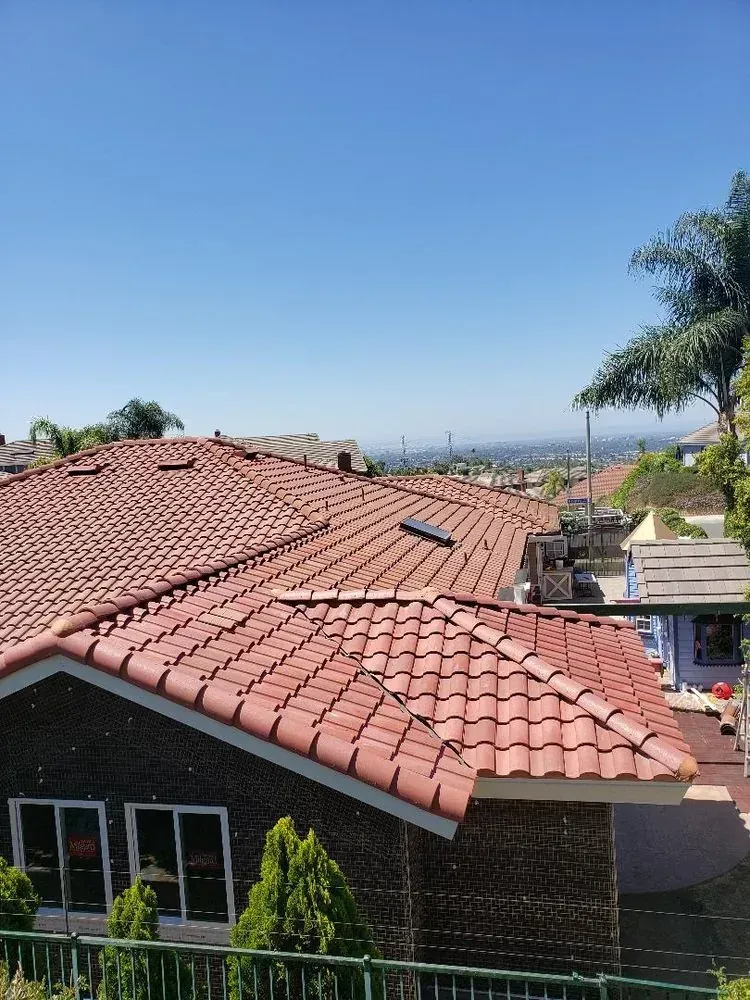 A view of a house with a red tile roof and dark brick walls, with a distant landscape under a clear blue sky.